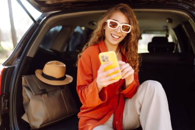 Güzel bir kadın açık havada bir arabanın bagajında oturuyor ve akıllı telefon kullanıyor. Seyahat, hafta sonu, doğa, rahatlama ve yaşam tarzı konsepti. Bloglama, iletişim.