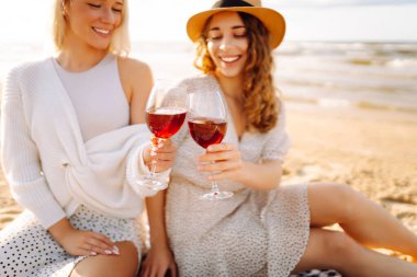 İki kız arkadaş kırmızı şarapla birlikte oturuyor, bir yaz öğleden sonra sahilde sohbet ediyorlar. Moda tarzı. Piknik konsepti