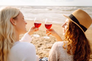 İki kız arkadaş kırmızı şarapla birlikte oturuyor, bir yaz öğleden sonra sahilde sohbet ediyorlar. Moda tarzı. Piknik konsepti