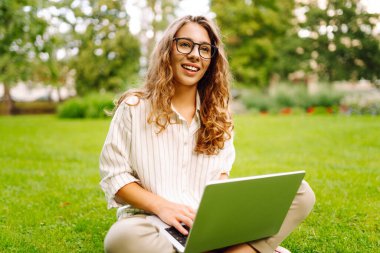 Yeşil bir açıklıkta dizüstü bilgisayarla oturan başarılı bir Avrupalı kadın. Bağımsız, Çevrimiçi, teknoloji konsepti.