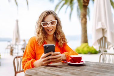Kafede kahve içerken cep telefonu kullanan mutlu güzel bir kadın. İnsanlar, moda, yaşam tarzı, seyahat ve tatil kavramı.