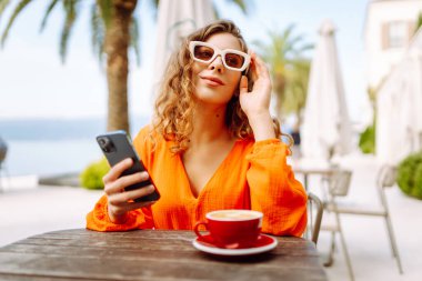 Kafede kahve içerken cep telefonu kullanan mutlu güzel bir kadın. İnsanlar, moda, yaşam tarzı, seyahat ve tatil kavramı.