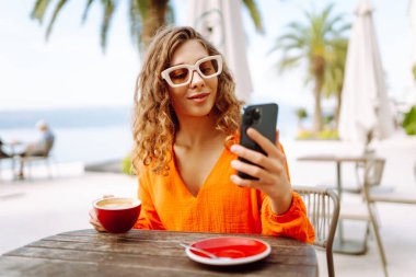 Kafede kahve içerken cep telefonu kullanan mutlu güzel bir kadın. İnsanlar, moda, yaşam tarzı, seyahat ve tatil kavramı.