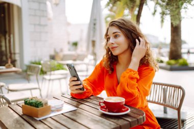 Kafede kahve içerken cep telefonu kullanan mutlu güzel bir kadın. İnsanlar, moda, yaşam tarzı, seyahat ve tatil kavramı.