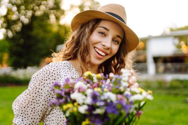 Bir buket kır çiçeğini elinde tutan şık bir yaz elbisesi ve şapkası olan genç ve güzel bir kadın. Yaz zamanı. Moda Baharı.