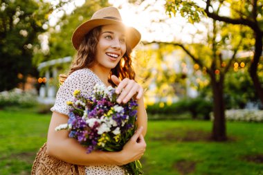 Bir buket kır çiçeğini elinde tutan şık bir yaz elbisesi ve şapkası olan genç ve güzel bir kadın. Yaz zamanı. Moda Baharı.