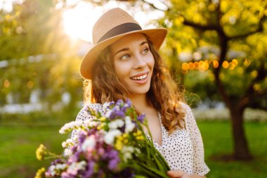 Bir buket kır çiçeğini elinde tutan şık bir yaz elbisesi ve şapkası olan genç ve güzel bir kadın. Yaz zamanı. Moda Baharı.