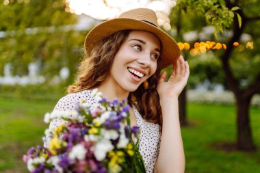 Bir buket kır çiçeğini elinde tutan şık bir yaz elbisesi ve şapkası olan genç ve güzel bir kadın. Yaz zamanı. Moda Baharı.