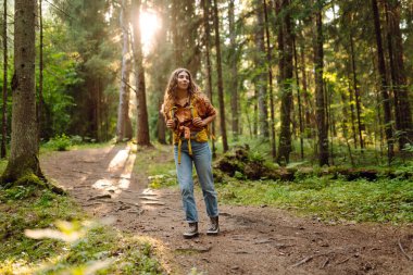 Ormanda ağaçların arasında yürürken sırt çantası taşıyan genç bir kadın. Yürüyüş, doğa, aktif yaşam tarzı