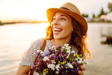 Beyaz elbiseli, elinde bir buket kır çiçeğiyle gün batımında yürüyen mutlu genç kız. Düğünün kırsal tarzı.