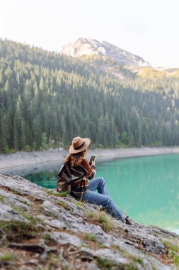 Sırt çantalı ve sıcak giysili genç bir bayan gezgin güzel bir gölün kenarındaki kayanın üzerinde duruyor, gülümsüyor ve telefonunu kullanıyor. Avrupa seyahati. Yaşam tarzı, tatil, turizm, doğa, aktif hayat. Günlük tutma