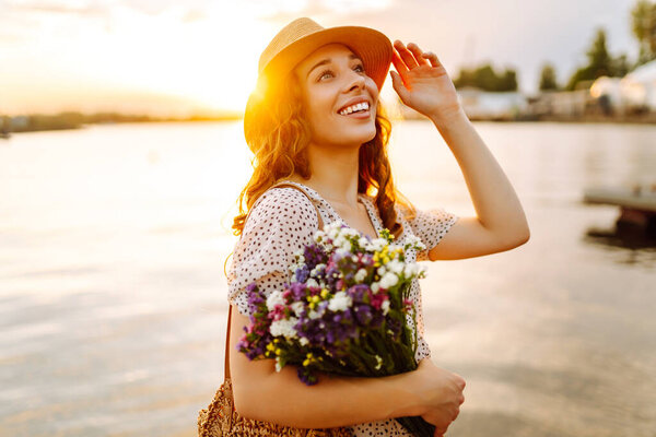 Счастливая молодая девушка в белом платье с букетом диких цветов гуляет по закату. Сельский стиль свадьбы