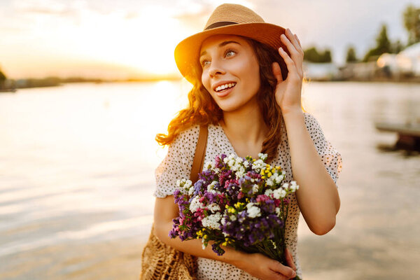 Счастливая молодая девушка в белом платье с букетом диких цветов гуляет по закату. Сельский стиль свадьбы