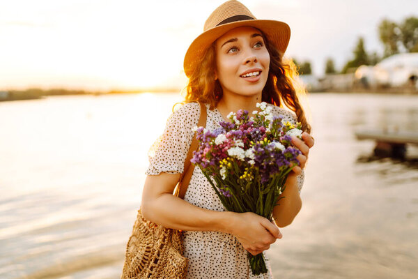 Счастливая молодая девушка в белом платье с букетом диких цветов гуляет по закату. Сельский стиль свадьбы