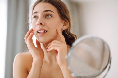 Güzel bir kadın aynaya bakıyor, eliyle yüzüne dokunuyor, sivilceleri ve pigmentasyonu inceliyor. Dermatoloji, kozmetoloji, cilt bakımı. Doğal cilt