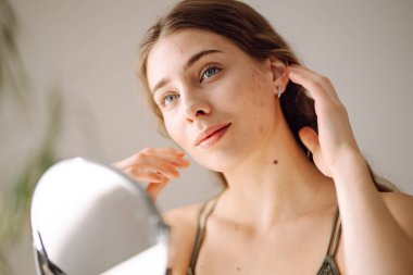 Güzel bir kadın aynaya bakıyor, eliyle yüzüne dokunuyor, sivilceleri ve pigmentasyonu inceliyor. Dermatoloji, kozmetoloji, cilt bakımı. Doğal cilt