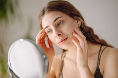 Güzel bir kadın aynaya bakıyor, eliyle yüzüne dokunuyor, sivilceleri ve pigmentasyonu inceliyor. Dermatoloji, kozmetoloji, cilt bakımı. Doğal cilt