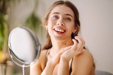 Güzel bir kadın aynaya bakıyor, eliyle yüzüne dokunuyor, sivilceleri ve pigmentasyonu inceliyor. Dermatoloji, kozmetoloji, cilt bakımı. Doğal cilt