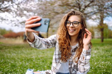 Parktaki piknikte ekose üzerinde otururken selfie çeken güzel, mutlu genç bir kadın. Bloglama, iletişim, rahatlama, hafta sonu