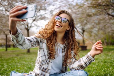 Parktaki piknikte ekose üzerinde otururken selfie çeken güzel, mutlu genç bir kadın. Bloglama, iletişim, rahatlama, hafta sonu