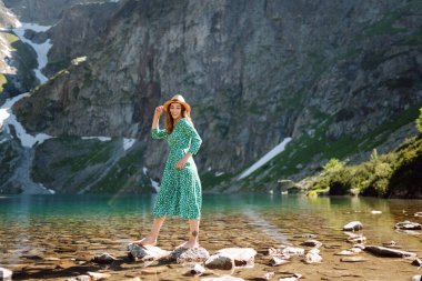 Genç bir kadın güneş ışınlarıyla dağ gölü manzarasının keyfini çıkarıyor. Dağlarda gezen şık elbiseli güzel bir kadın. Macera konsepti, turist, tatil.