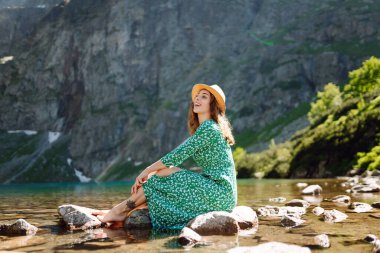 Genç bir kadın güneş ışınlarıyla dağ gölü manzarasının keyfini çıkarıyor. Dağlarda gezen şık elbiseli güzel bir kadın. Macera konsepti, turist, tatil.