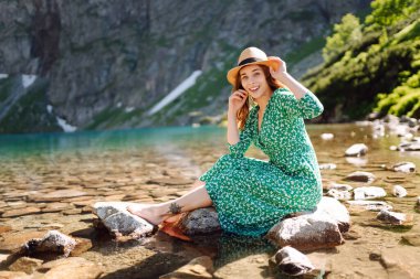 Genç bir kadın güneş ışınlarıyla dağ gölü manzarasının keyfini çıkarıyor. Dağlarda gezen şık elbiseli güzel bir kadın. Macera konsepti, turist, tatil.