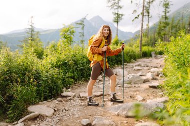 Sırt çantalı ve yürüyüş direkleri olan kadın yürüyüşçü arka planda dağlarla kayalık yola tırmanıyor. Güneşli bir günde manzaranın keyfini çıkaran sportif bir kadın. Yürüyüş konsepti. Etkin yaşam biçimi.