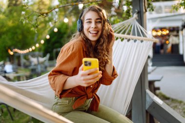 Kablosuz kulaklık ve telefonla müzik dinleyen güzel bir kadının portresi. Genç kadın bir hamakta dinleniyor ve parkta doğanın tadını çıkarıyor. Müzik kavramı, blog yazma.