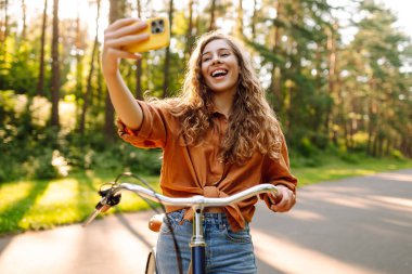 Neşeli bir kadın bisiklet sürüyor ve güneşli parkta selfie çekiyor. Güzel bir kadın dışarıda telefonla vakit geçiriyor. Selfie, boş zaman konsepti. Etkin yaşam biçimi.