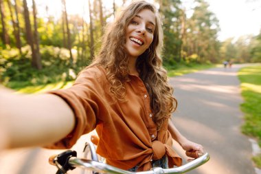 Neşeli bir kadın bisiklet sürüyor ve güneşli parkta selfie çekiyor. Güzel bir kadın dışarıda telefonla vakit geçiriyor. Selfie, boş zaman konsepti. Etkin yaşam biçimi.
