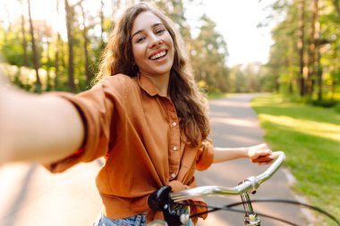 Neşeli bir kadın bisiklet sürüyor ve güneşli parkta selfie çekiyor. Güzel bir kadın dışarıda telefonla vakit geçiriyor. Selfie, boş zaman konsepti. Etkin yaşam biçimi.