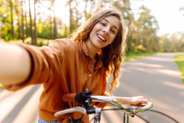 Neşeli bir kadın bisiklet sürüyor ve güneşli parkta selfie çekiyor. Güzel bir kadın dışarıda telefonla vakit geçiriyor. Selfie, boş zaman konsepti. Etkin yaşam biçimi.