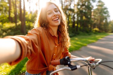 Neşeli bir kadın bisiklet sürüyor ve güneşli parkta selfie çekiyor. Güzel bir kadın dışarıda telefonla vakit geçiriyor. Selfie, boş zaman konsepti. Etkin yaşam biçimi.