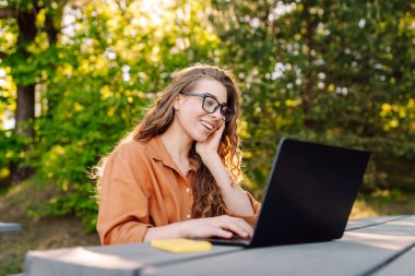 Parktaki ahşap bir masada dizüstü bilgisayarını kullanan neşeli bir kadın. Parktaki su kenarında çalışan serbest çalışan genç bir bayan. Boş zaman, iş konsepti.