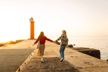 Deniz kenarında deniz feneri manzaralı iki genç bayan arkadaş eğleniyor. Güzel kadınlar gün batımında birlikte vakit geçirirler. Yürüme, eğlence, arkadaşlık kavramı.