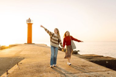Deniz kenarında deniz feneri manzaralı iki genç bayan arkadaş eğleniyor. Güzel kadınlar gün batımında birlikte vakit geçirirler. Yürüme, eğlence, arkadaşlık kavramı.