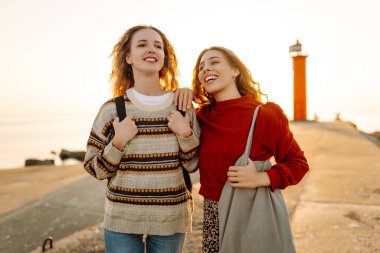 Deniz kenarında deniz feneri manzaralı iki genç bayan arkadaş eğleniyor. Güzel kadınlar gün batımında birlikte vakit geçirirler. Yürüme, eğlence, arkadaşlık kavramı.