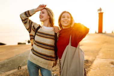 Deniz kenarında deniz feneri manzaralı iki genç bayan arkadaş eğleniyor. Güzel kadınlar gün batımında birlikte vakit geçirirler. Yürüme, eğlence, arkadaşlık kavramı.