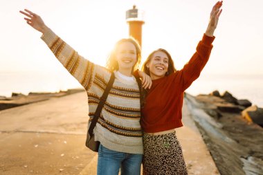 Deniz kenarında deniz feneri manzaralı iki genç bayan arkadaş eğleniyor. Güzel kadınlar gün batımında birlikte vakit geçirirler. Yürüme, eğlence, arkadaşlık kavramı.