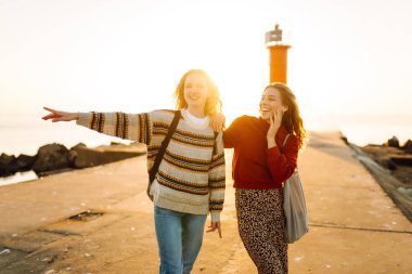 Deniz kenarında deniz feneri manzaralı iki genç bayan arkadaş eğleniyor. Güzel kadınlar gün batımında birlikte vakit geçirirler. Yürüme, eğlence, arkadaşlık kavramı.