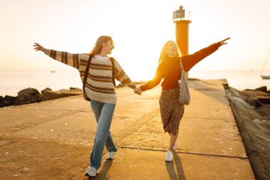 Deniz kenarında deniz feneri manzaralı iki genç bayan arkadaş eğleniyor. Güzel kadınlar gün batımında birlikte vakit geçirirler. Yürüme, eğlence, arkadaşlık kavramı.