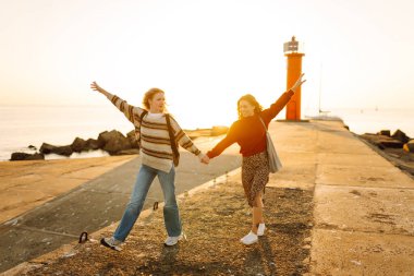 Deniz kenarında deniz feneri manzaralı iki genç bayan arkadaş eğleniyor. Güzel kadınlar gün batımında birlikte vakit geçirirler. Yürüme, eğlence, arkadaşlık kavramı.