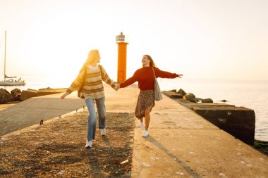 Deniz kenarında deniz feneri manzaralı iki genç bayan arkadaş eğleniyor. Güzel kadınlar gün batımında birlikte vakit geçirirler. Yürüme, eğlence, arkadaşlık kavramı.
