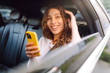 Kıvırcık saçlı, elinde telefonla arabanın arka koltuğunda gülümseyen genç bir kadın. Gülümseyen kadın taksiden zevk alıyor. Teknoloji ve iş konsepti.