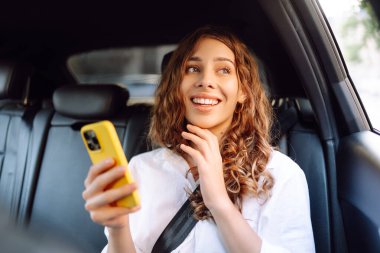 Kıvırcık saçlı, elinde telefonla arabanın arka koltuğunda gülümseyen genç bir kadın. Gülümseyen kadın taksiden zevk alıyor. Teknoloji ve iş konsepti.