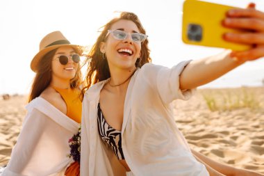 İki genç arkadaş deniz kenarındaki sahilde selfie çekiyorlar. Güzel kadınlar gün batımının tadını çıkarıyorlar. Arkadaşlık konsepti, rahatlama, blog yazma. Yaşam biçimi.