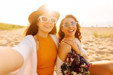 İki genç arkadaş deniz kenarındaki sahilde selfie çekiyorlar. Güzel kadınlar gün batımının tadını çıkarıyorlar. Arkadaşlık konsepti, rahatlama, blog yazma. Yaşam biçimi.
