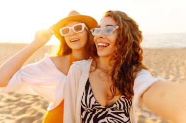 İki genç arkadaş deniz kenarındaki sahilde selfie çekiyorlar. Güzel kadınlar gün batımının tadını çıkarıyorlar. Arkadaşlık konsepti, rahatlama, blog yazma. Yaşam biçimi.
