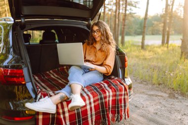 Güneşli bir günde dizüstü bilgisayarla bir arabanın bagajında oturan genç bir kadın. Modern bir kadın dışarıda dizüstü bilgisayarda çalışıyor. Arabayla, blogla, uzaktan çalışma kavramı.